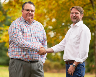 Two men shake hands in front of green and yellow trees