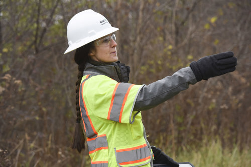 A woman in a hard hat and construction vest points to the right in front of trees.