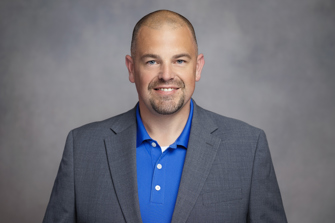 A headshot of a man in a gray suit jacket and bright blue button-up shirt against a gray background.